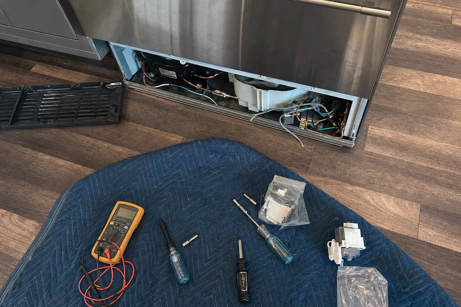 Base grille off an undercounter unit with the compressor exposed, multimeter, drivers and a bagged spare part on a work mat